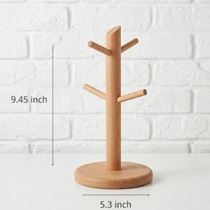 May include: A wooden mug tree with a round base and four horizontal pegs for hanging mugs. The tree is 9.45 inches tall and 5.3 inches wide. The natural wood finish complements the white brick background.