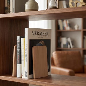 May include: Wooden bookshelf with books and bookends. The books include titles like "Vermeer" and "Johannes Vermeer". The bookends are rectangular and made of light-colored wood. The shelf is made of dark wood.