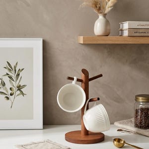 May include: A wooden mug tree with two white ceramic mugs hanging from it. The mug tree is made of dark brown wood and has a round base. A framed botanical print and a jar of coffee beans are also visible.