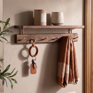 Könnte beinhalten: Eine Wandablage aus Holz mit Haken. Ein braun-weiß karierter Schal hängt an einem Haken, und ein Leder-Schlüsselanhänger mit Schlüsseln hängt an einem anderen. Ein zylindrischer Behälter und eine strukturierte Vase stehen auf dem Regal.