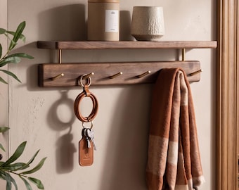 Puede incluir: Estante de pared de madera con ganchos. Una bufanda marrón y blanca cuelga de un gancho, y un llavero de cuero con llaves cuelga de otro. Un recipiente cilíndrico y un jarrón texturizado se encuentran en el estante.