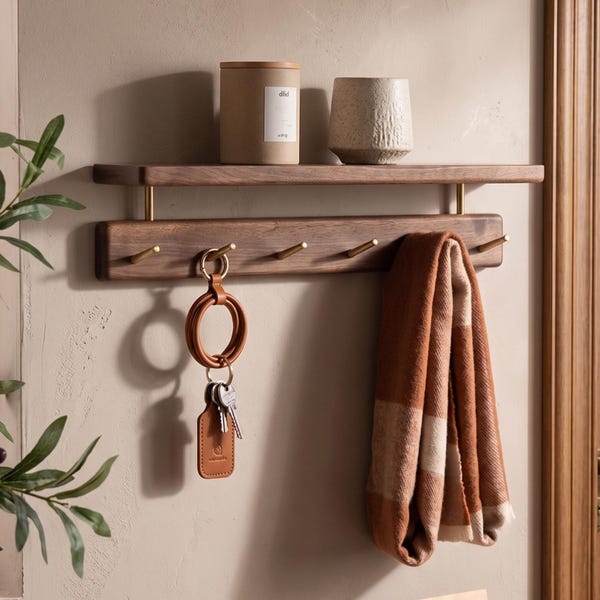 May include: A wall-mounted dark wood shelf with a lower rail featuring five brass hooks. A tan leather keychain with keys and a brown and white plaid scarf hang from the hooks. A cylindrical container and a textured beige cup sit on the shelf.