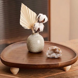 May include: A round wooden tray with three light-colored feet holds a small speckled ceramic vase containing a dried palm leaf and cotton stems, alongside a halved geode with visible crystals, creating a balanced display of natural textures.