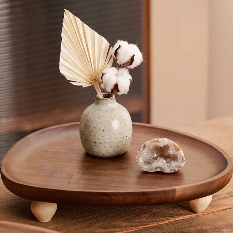 May include: A round wooden tray with three light-colored feet holds a small speckled ceramic vase containing a dried palm leaf and cotton stems, alongside a halved geode with visible crystals, creating a balanced display of natural textures.