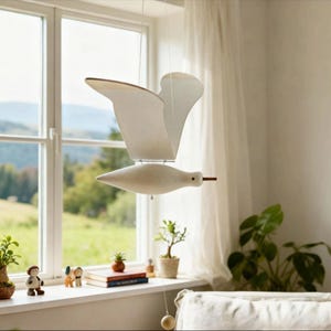 May include: A hanging mobile featuring a stylized white bird with outstretched wings suspended by thin wires. The bird has a smooth, matte finish and a small dark eye. A thin string hangs from the bird's body, ending in a small wooden sphere.