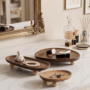 May include: Three dark brown wooden trays with light-colored spherical feet. The long rectangular tray holds a white bust figurine and a beaded necklace. The large oval tray holds a wristwatch and a black case. The small oval tray holds rings.