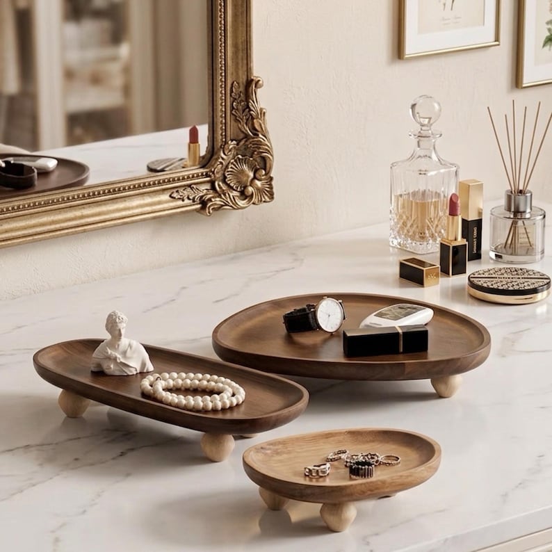 May include: Three dark brown wooden trays with light-colored spherical feet. The long rectangular tray holds a white bust figurine and a beaded necklace. The large oval tray holds a wristwatch and a black case. The small oval tray holds rings.