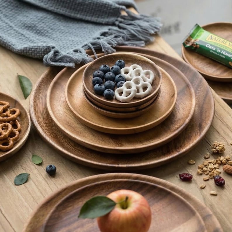 May include: Wooden serving plates of various sizes are stacked, some holding blueberries and pretzels. A green and white granola bar is visible in the background. The plates are on a light brown wooden surface, with a gray fringed cloth in the background.