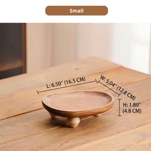 May include: A small oval wooden tray with three short rounded legs. The tray features a natural wood grain finish and measures 6.50 inches in length, 5.04 inches in width, and 1.89 inches in height. The text "Small" is displayed above the tray.