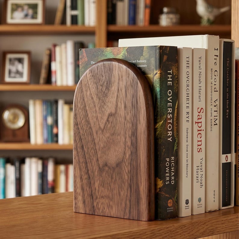 May include: A wooden arched bookend in a warm brown tone stands next to a row of books. The bookend's smooth, curved design complements the spines of the books, creating a cohesive look. The books include titles like "Sapiens" and "The Overstory".