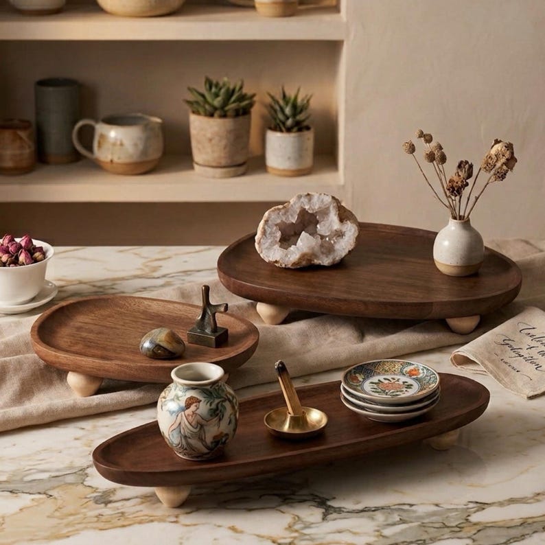 May include: Three oval wooden trays with small rounded feet, featuring a smooth dark brown finish. The trays hold various decorative items including a geode, a small ceramic vase with a figure, a brass incense holder, and small patterned plates.