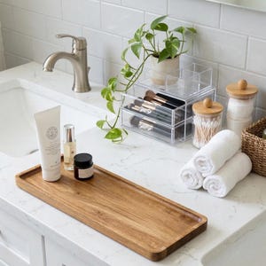 May include: A wooden rectangular tray holds skincare products, a small black jar, and a white tube. A clear acrylic organizer with makeup brushes sits behind a small potted plant. Rolled white towels and cotton swabs in a jar are also visible.