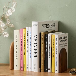 May include: A collection of hardcover books with various titles, including "Vermeer" and "The Story of Art," arranged between two wooden bookends. The books are in different colors, with titles in English. The background is a light green wall.