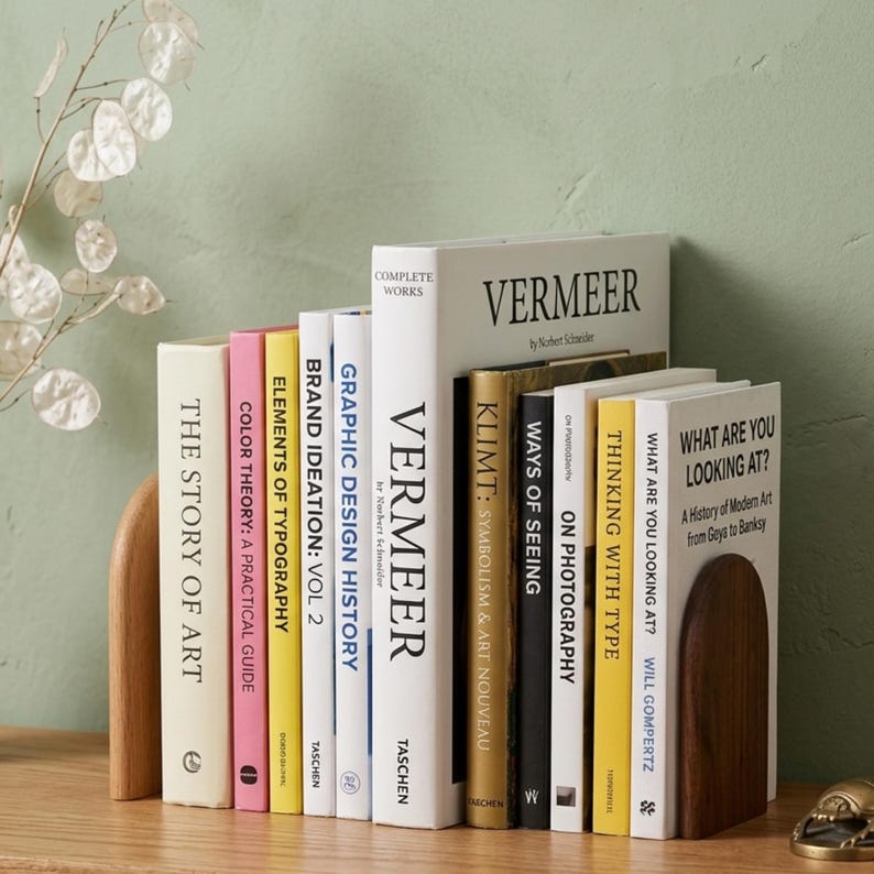 May include: A collection of hardcover books with various titles, including "Vermeer" and "The Story of Art," arranged between two wooden bookends. The books are in different colors, with titles in English. The background is a light green wall.