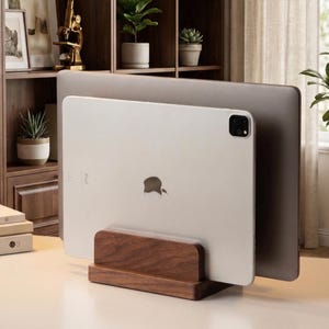 May include: A wooden desk organizer holds a silver iPad and a laptop. The organizer is made of dark brown wood and has a rectangular base. The iPad has the Apple logo on the back. The background includes a wooden shelf with plants.