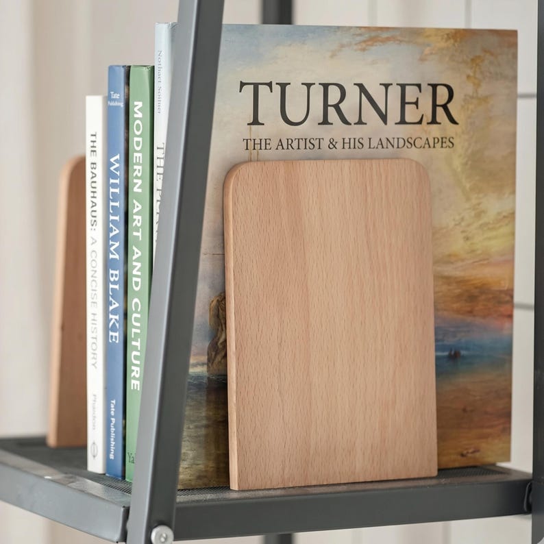 May include: A set of wooden bookends holding a collection of books on a metal shelf. The books include titles like "TURNER", "WILLIAM BLAKE", and "MODERN ART AND CULTURE". The bookends are rectangular and made of light-colored wood.