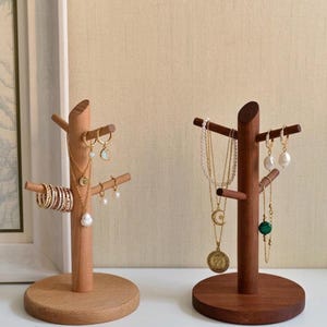 May include: Two wooden jewelry display stands. One is light-colored, the other is dark. The light-colored stand holds gold rings, earrings, and necklaces. The dark-colored stand holds necklaces and earrings. Both stands have a tree-like design.