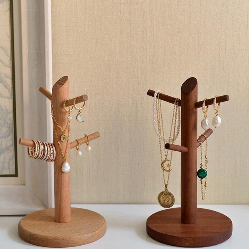 May include: Two wooden jewelry display stands. One is light-colored, the other is dark. The light-colored stand holds gold rings, earrings, and necklaces. The dark-colored stand holds necklaces and earrings. Both stands have a tree-like design.
