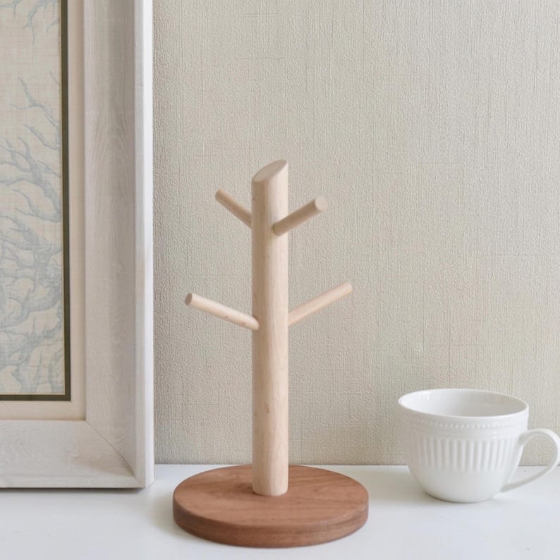 May include: A light-colored wooden mug tree with four branches, mounted on a round, dark brown base. A white teacup with a handle sits to the right. A framed picture is on the left.