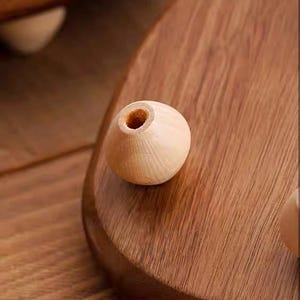 May include: A light-colored, spherical wooden bead with a central hole, resting on a smooth, polished wooden surface with a visible grain pattern. The bead has a natural, unfinished appearance and a rounded shape, contrasting with the dark wood.
