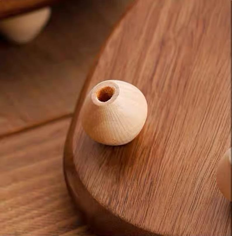 May include: A light-colored, spherical wooden bead with a central hole, resting on a smooth, polished wooden surface with a visible grain pattern. The bead has a natural, unfinished appearance and a rounded shape, contrasting with the dark wood.