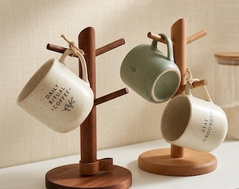 May include: Two wooden mug trees with round bases, each holding ceramic mugs. One tree holds a cream-colored mug with the text 'DAILY RITUAL COFFEE.' The other holds a sage green mug and a cream-colored mug with the text 'STAY GROUNDED.'