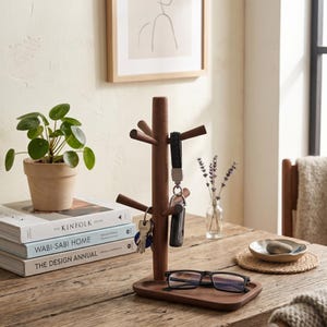 May include: A wooden key holder with a tree-like design, featuring multiple pegs for keys and a tray for small items. The key holder is made of dark brown wood and holds keys, a key fob, and eyeglasses. A small potted plant and books are also on the table.
