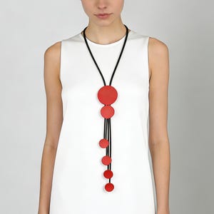 Può includere: Una lunga collana con pendenti circolari rossi di diverse dimensioni, sospesi a cordini neri. La collana ha un grande pendente circolare in alto, con cerchi più piccoli a cascata.