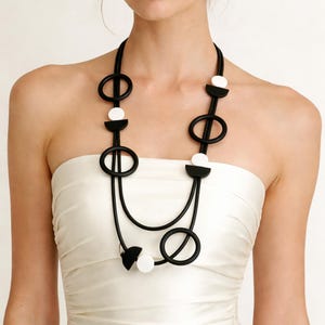 Può includere: Una collana geometrica in bianco e nero. La collana presenta una serie di forme ovali e a semicerchio nere, intervallate da perline bianche circolari. La collana è sospesa da un cordoncino nero, creando un design moderno e artistico. Un gioiello audace.