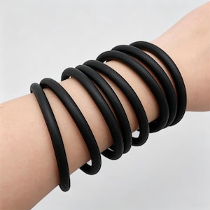 May include: A collection of black rubber bracelets are stacked on a wrist. The bracelets are round and have a smooth texture. The bracelets are a solid black color and appear to be made of a flexible material.