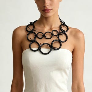 Può includere: Una collana nera audace con molteplici elementi circolari interconnessi. La collana è realizzata in un materiale scuro e viene indossata sopra un top bianco senza spalline. Il design incorpora nodi per collegare le forme circolari.