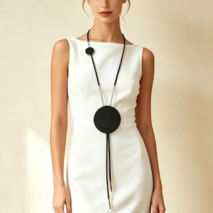 May include: A black necklace with a large circular pendant and a smaller circular accent. The necklace has a long, slender cord with metallic accents. The design is minimalist and modern, suitable for various outfits.