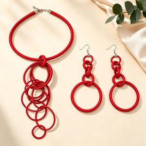 Può includere: Un set di gioielli rosso brillante composto da una collana girocollo e un paio di orecchini pendenti abbinati. La collana presenta una spessa fascia in corda con un gruppo di anelli circolari di varie dimensioni appesi al centro del design.