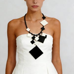 Può includere: Collana geometrica in bianco e nero con pendenti a forma di diamante. La collana, con un cordino nero, è indossata su un abito bianco senza spalline. Il design è moderno e audace, con un contrasto sorprendente.