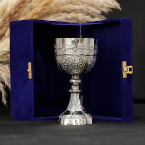 May include: A silver chalice with ornate floral detailing, presented inside a deep blue velvet box. The chalice has a detailed stem and a wide, decorative bowl. The box is open, revealing the chalice against a dark background.