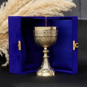 Puede incluir: Un cáliz dorado con intrincados diseños en relieve, presentado dentro de una caja de terciopelo azul oscuro abierta. La caja tiene cierres dorados. El cáliz es ornamentado, con un tallo y una copa detallados. El fondo incluye hierba ornamental seca.