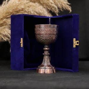 May include: An ornate copper-colored chalice with detailed floral patterns, presented inside a deep blue velvet box. The chalice has a tall stem and a wide bowl, showcasing intricate carvings. The box is open, revealing the chalice against a dark background.
