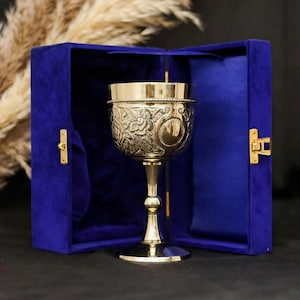 May include: A gold-colored chalice with intricate floral and scrollwork designs, presented in an open, deep blue velvet box. The chalice has a tall stem and a wide, detailed bowl. The box has gold-colored hardware and a soft, plush interior.