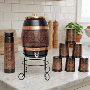 May include: A decorative water dispenser in black and copper, with a matching water bottle and a stack of cups. The dispenser has an ornate pattern and sits on a black metal stand. The cups and bottle have the same design.