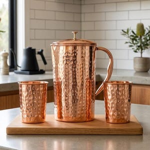 Könnte beinhalten: Ein Kupferkrug mit Deckel und Griff, zusammen mit zwei passenden Kupferbechern, alle mit gehammerter Textur. Das Set wird auf einem Holzservierbrett präsentiert.