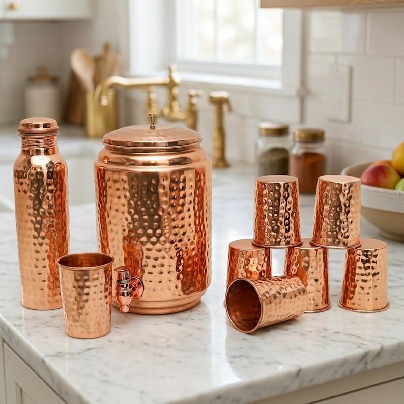 Puede incluir: Un conjunto de utensilios de cocina de cobre, que incluye una botella de agua, un gran dispensador de bebidas de cobre martillado con grifo y varias tazas de cobre martillado. Los art&iacute;culos tienen una superficie texturizada y un color marr&oacute;n rojizo. El fondo muestra una cocina.
