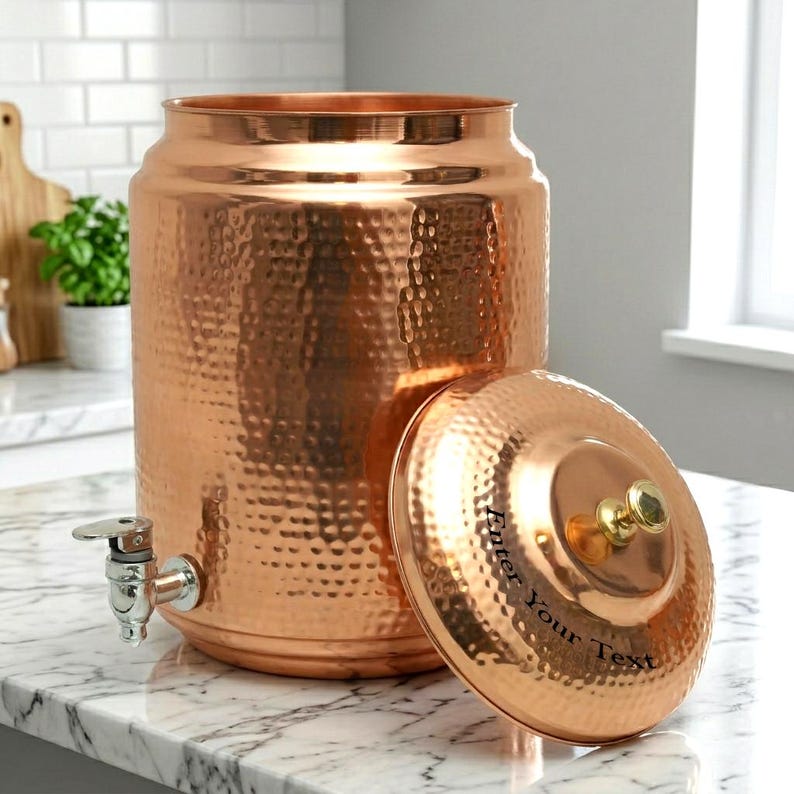 Puede incluir: Un dispensador de agua de color cobre con textura martillada. Tiene un grifo y una tapa con el texto "Enter Your Text". La tapa tiene un pomo dorado. El dispensador est&aacute; sobre una encimera de m&aacute;rmol.