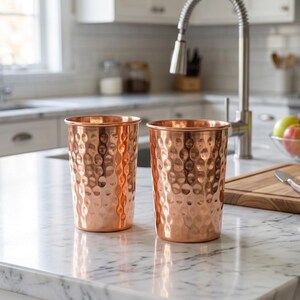May include: Two copper-colored drinking glasses with a hammered texture are displayed on a marble countertop. The glasses have a cylindrical shape with a slightly flared rim. The kitchen setting includes a stainless steel faucet and a cutting board.