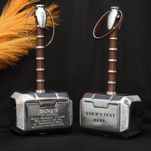 May include: Two silver-colored Thor's hammers with brown handles and silver accents. One hammer has the text "Thor" and a quote. The other hammer has the text "YOUR'S TEXT HERE". The hammers have a loop at the top.