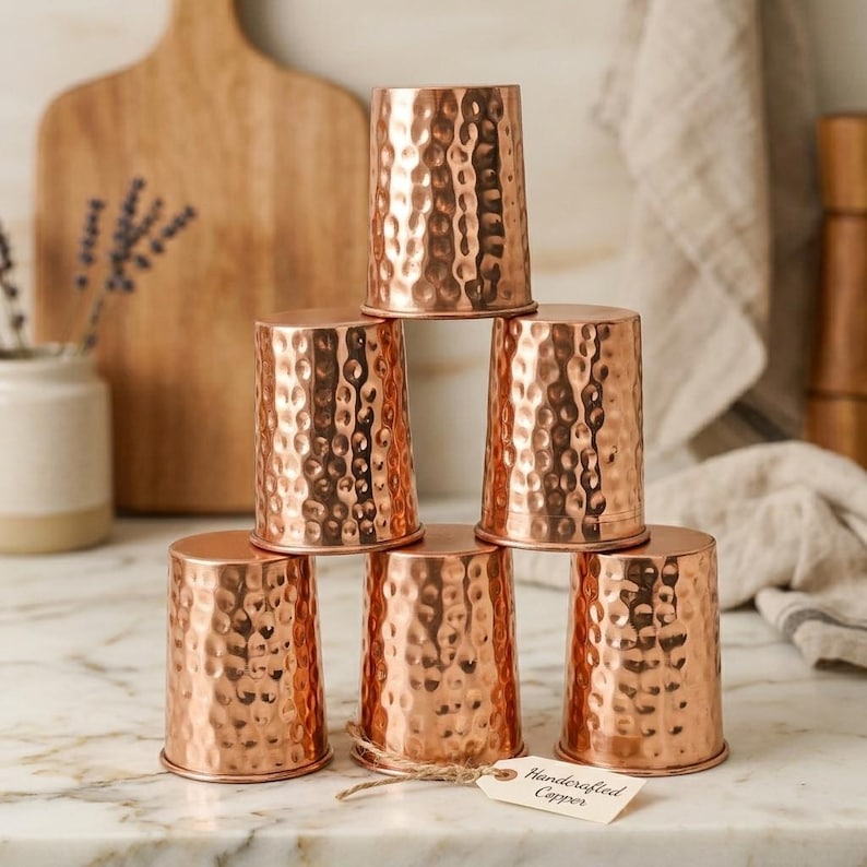 Puede incluir: Seis tazas de cobre con textura martillada, apiladas. Cada taza es cil&iacute;ndrica con un borde ensanchado. Una etiqueta con la inscripci&oacute;n "Handcrafted Copper" est&aacute; adjunta. Las tazas est&aacute;n sobre una superficie blanca, con una tabla de madera al fondo.