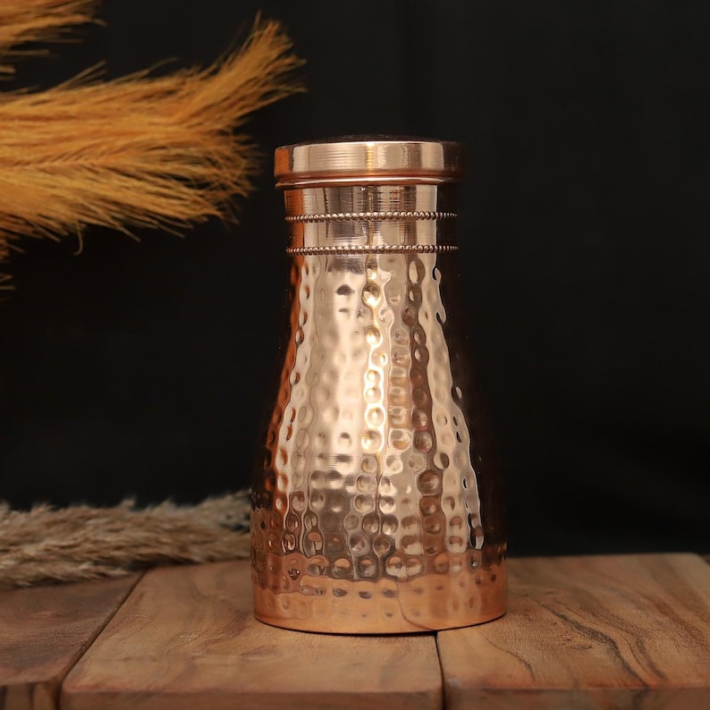 May include: A copper-colored water bottle with a hammered texture. The bottle has a cylindrical neck with decorative bands and a flat lid. The bottle is displayed on a wooden surface with dried ornamental grass in the background.