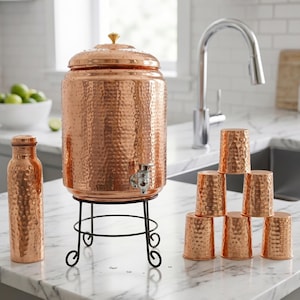 Puede incluir: Dispensador de agua de cobre con textura martillada, grifo y tapa con pomo dorado. Se apoya sobre un soporte de metal negro. Tambi&eacute;n hay una botella de agua y una pila de tazas de cobre a juego.