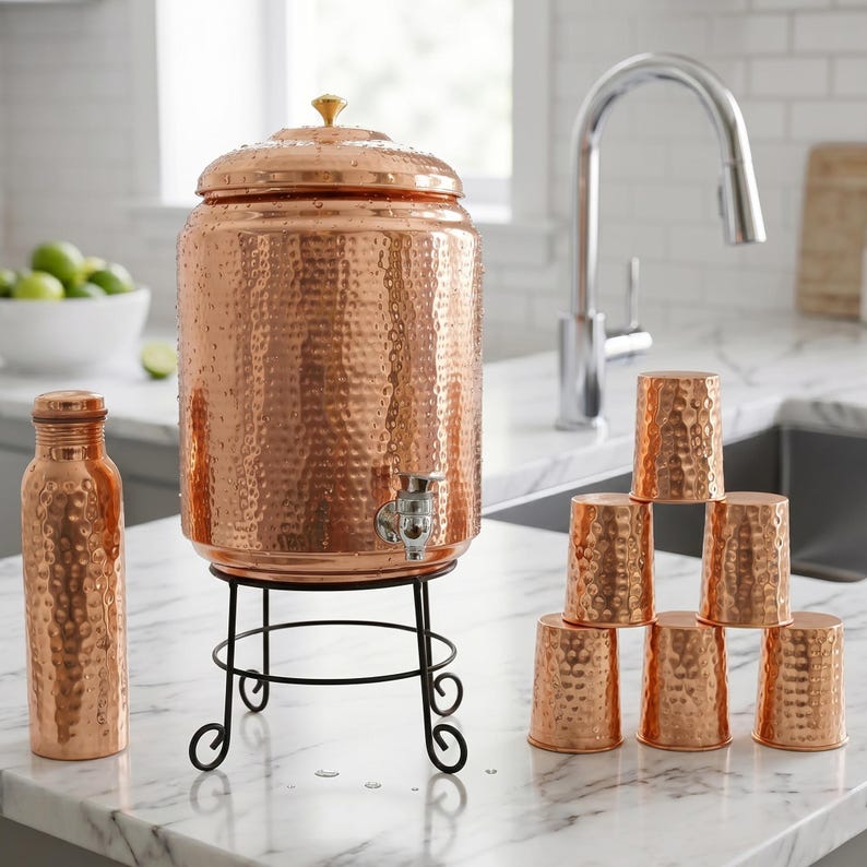 Puede incluir: Dispensador de agua de cobre con textura martillada, grifo y tapa con pomo dorado. Se apoya sobre un soporte de metal negro. Tambi&eacute;n hay una botella de agua y una pila de tazas de cobre a juego.