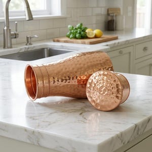May include: A copper-colored water pitcher with a hammered texture and a matching lid. The pitcher is lying on its side, with the lid next to it. The kitchen countertop is white marble.