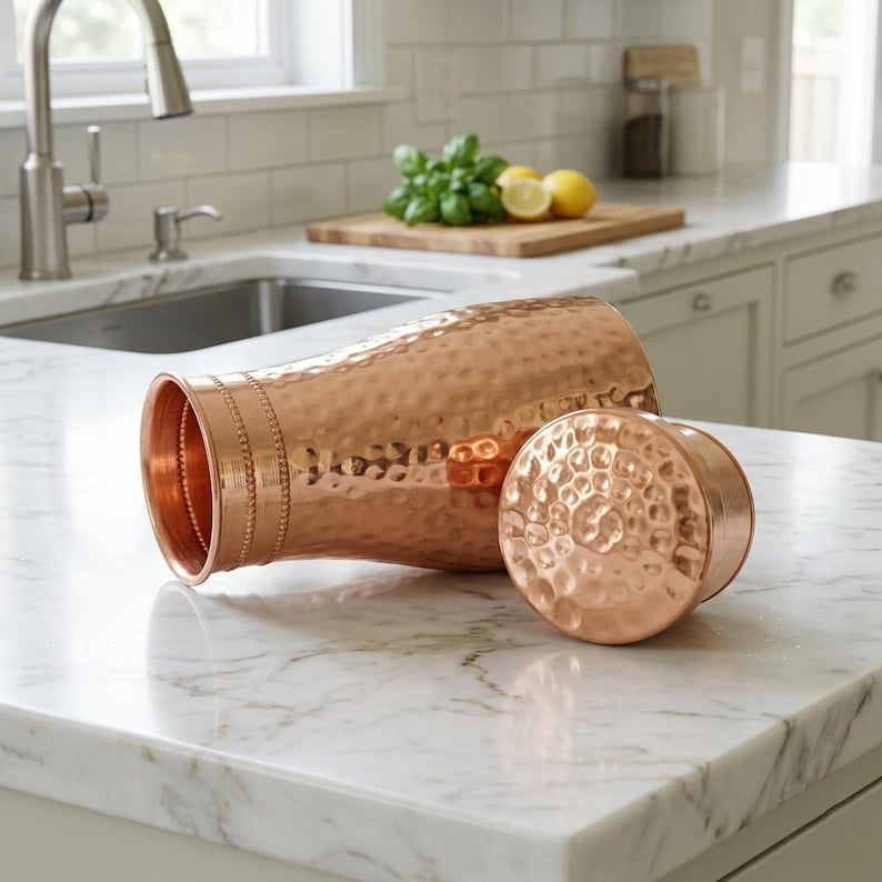 May include: A copper-colored water pitcher with a hammered texture and a matching lid. The pitcher is lying on its side, with the lid next to it. The kitchen countertop is white marble.
